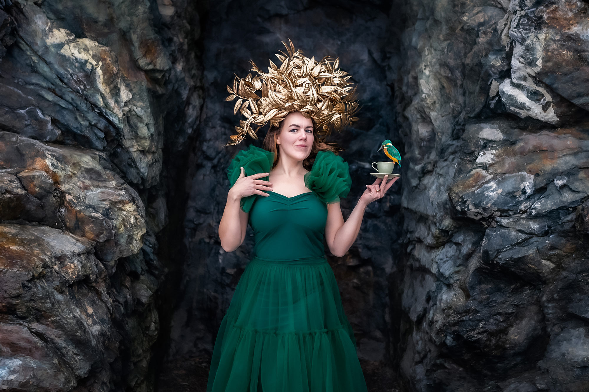 En kvinna i grön klänning och med en gyllene bladkrona på huvudet står i en klippöppning. Kvinnan håller en grön tekopp och på den sitter en liten grön och orange fågel.