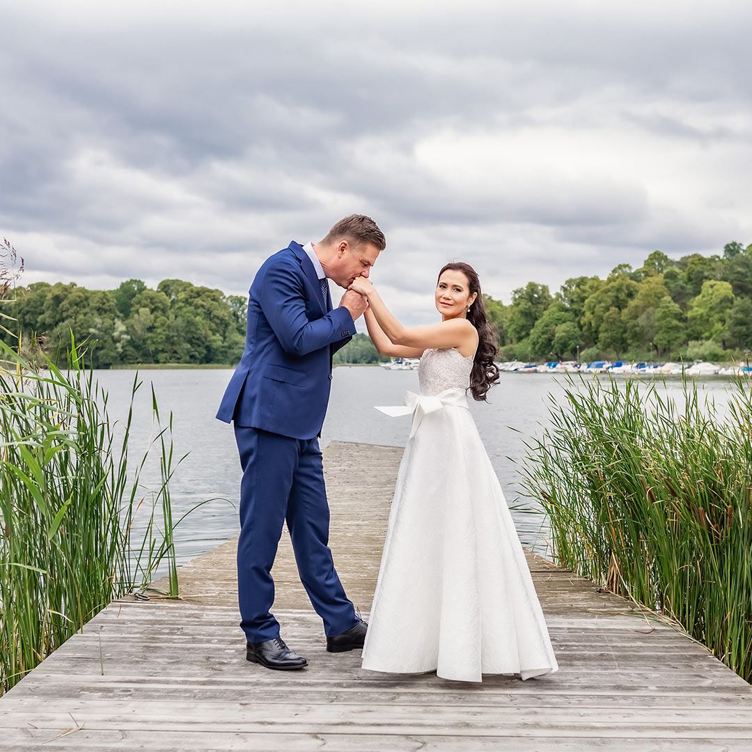 Brudpar står på en brygga vid vattnet och omgivna av vass. Mannen pussar kvinnan på händerna och kvinnan tittar in i bröllopsfotograf Linda Holmkratz kamera.