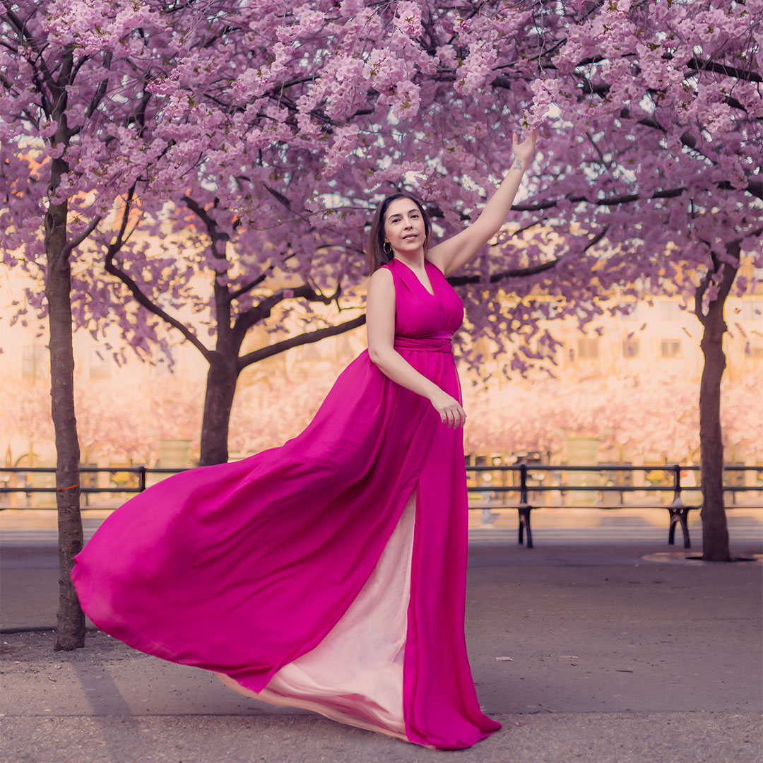 En kvinna i lång rosa klänning sträcker sig mot blomstrande körsbärsträd.