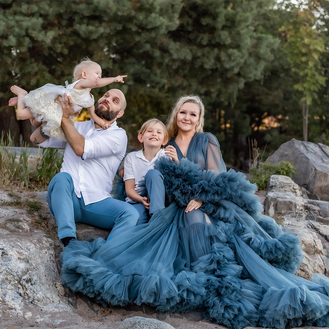 En familj med snygga blå kläder sitter på klippor. I bakgrunden syns barrträd. Pappan lyfter upp den lilla flickan och hela familjen ler in i kameran.