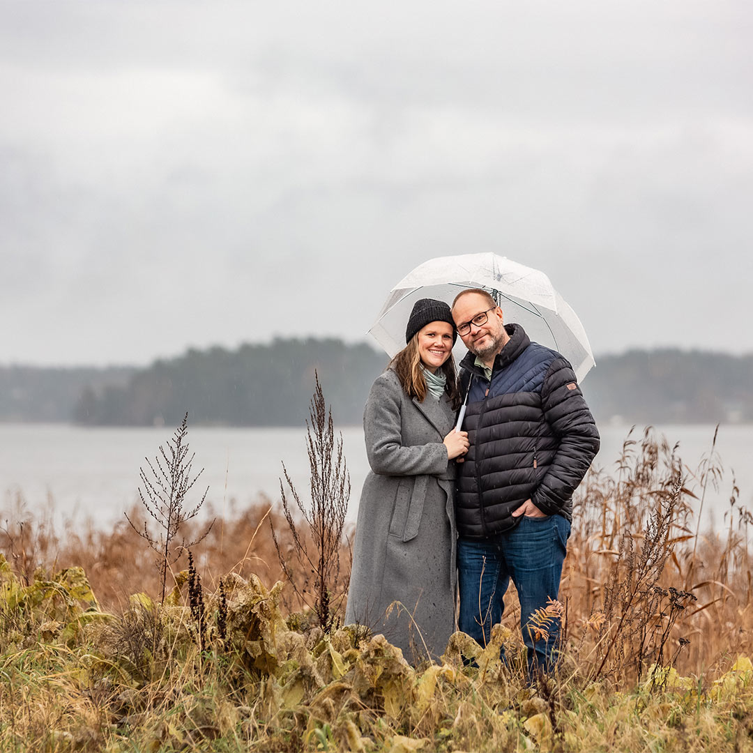 Ett par står i ett regndisigt landskap bland höga gräs. De håller i ett genomskinligt paraply och i bakgrunden syns havet.