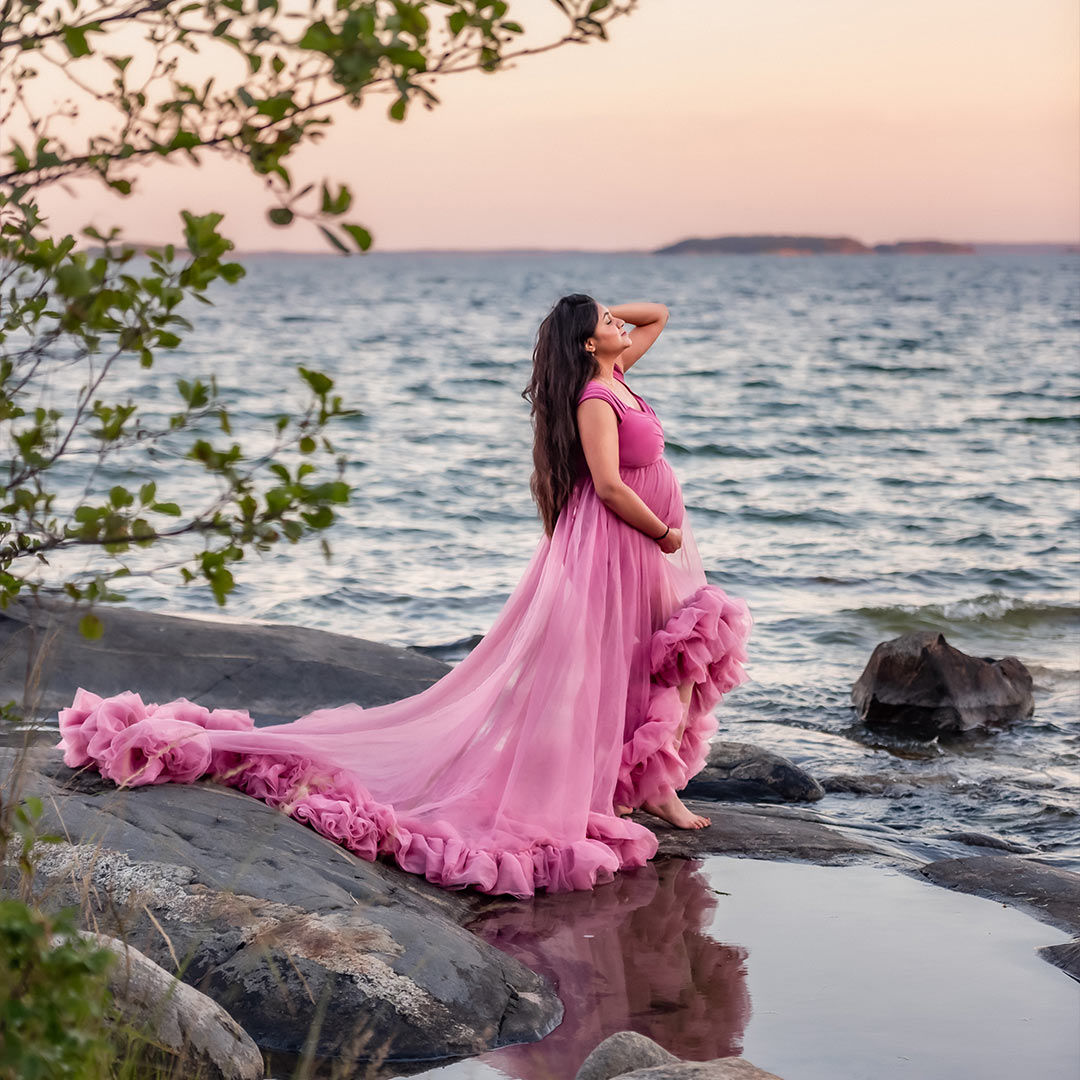 Gravidfotografering en sommarkväll i skärgården. I förgrunden syns grenar med gröna blad och i bakgrunden är en rosa himmel över vågigt hav. Den gravida kvinnan står på klipporna med en pöl vatten där hon speglas. Hon har på sig en rosa gravidklänning med mycket tyll.
