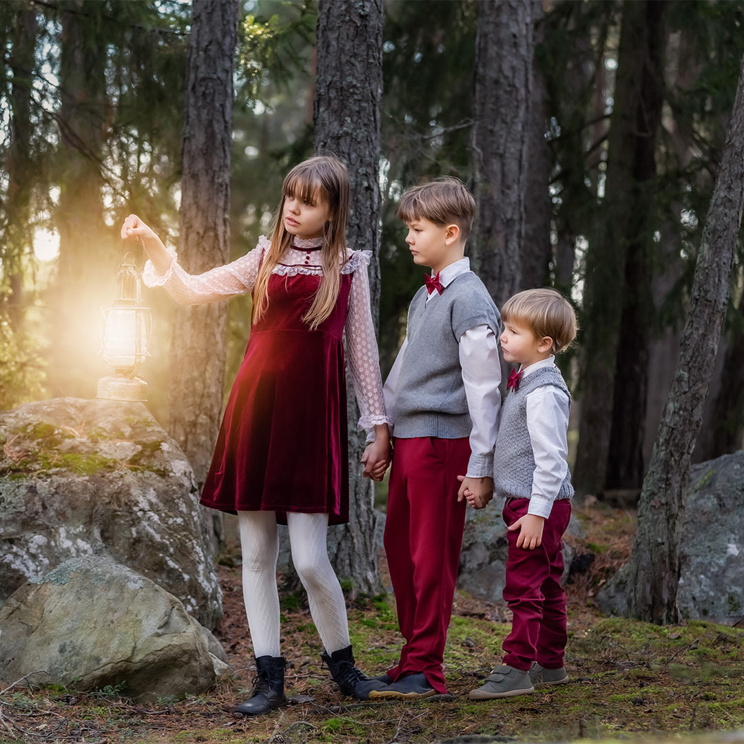 Tre syskon i röda julkläder går på rad med en lysande lykta i en barrskog med mossiga stora stenar.
