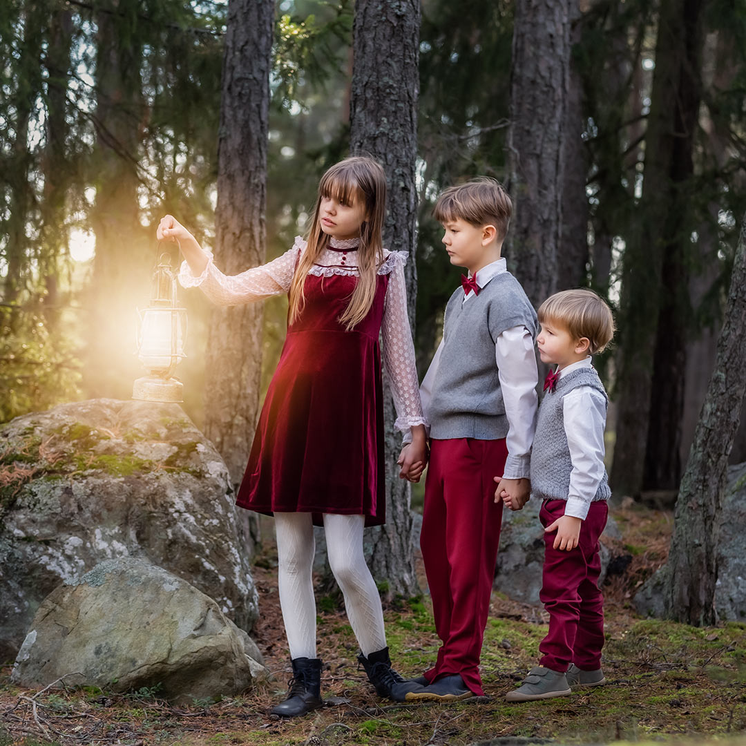 Tre barn i röda finkläder går hand i hand i en barrskog. Den äldsta flickan går längst fram och håller en gammal lykta som lyser upp deras väg.