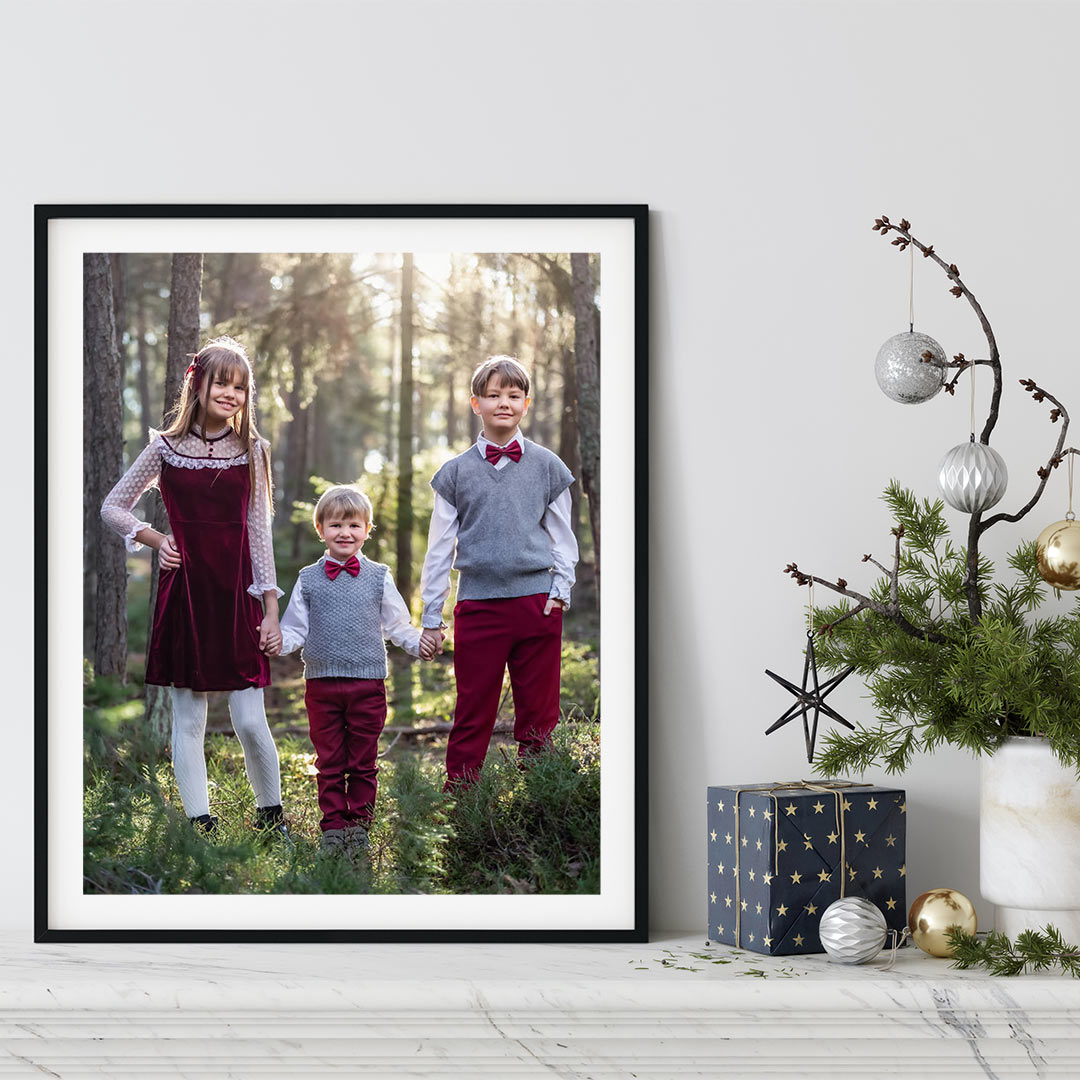 En tavelram står på en bänk bredvid juldekorationer. I ramen är ett foto på tre barn i röda finkläder.
