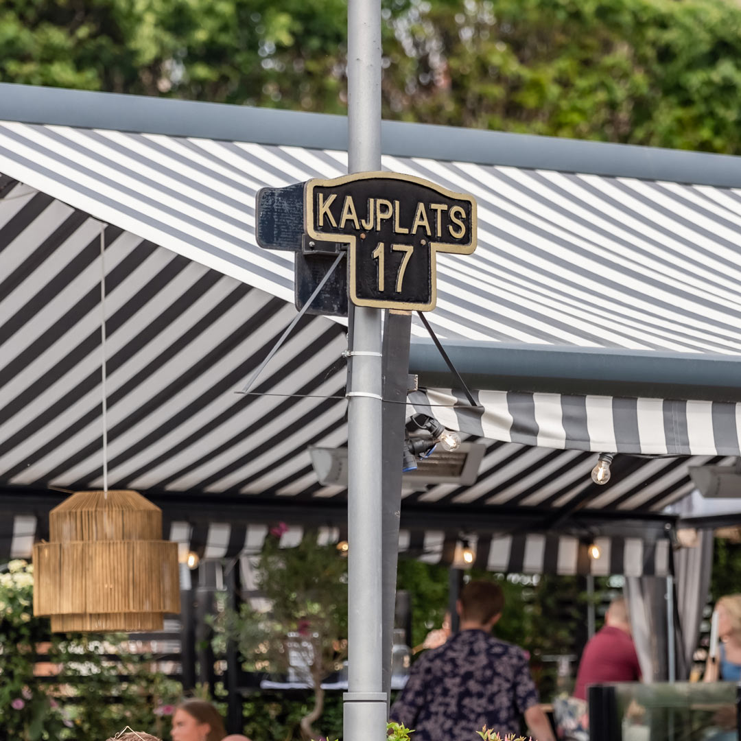 En skylt som säger Kajplats 17. Skylten står på en kaj och i bakgrunden syns en restaurang.