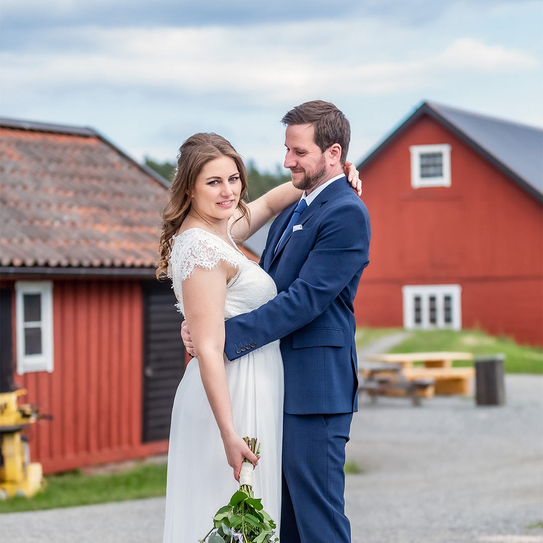 Ett brudpar står på en bondgård och håller om varandra. Kvinnan tittar in i kameran medan mannen tittar på kvinnan. I bakgrunden syns röda ladugårdar.