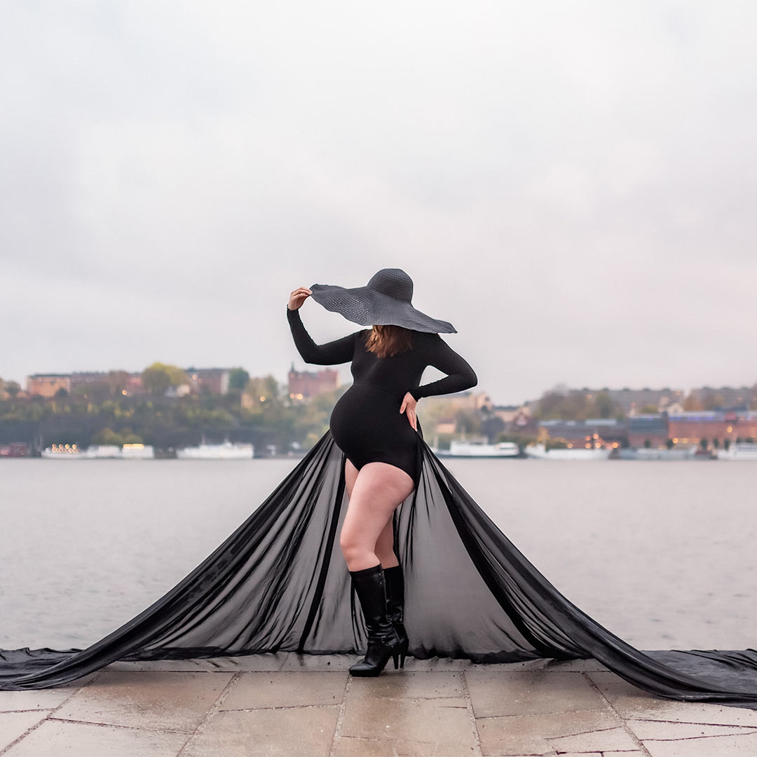 En gravid kvinna har på sig en svart body med ett långt utslaget släp och en gigantisk svart hatt. I bakgrunden syns vatten och i horisonten en stad. Det är gråmulet väder.