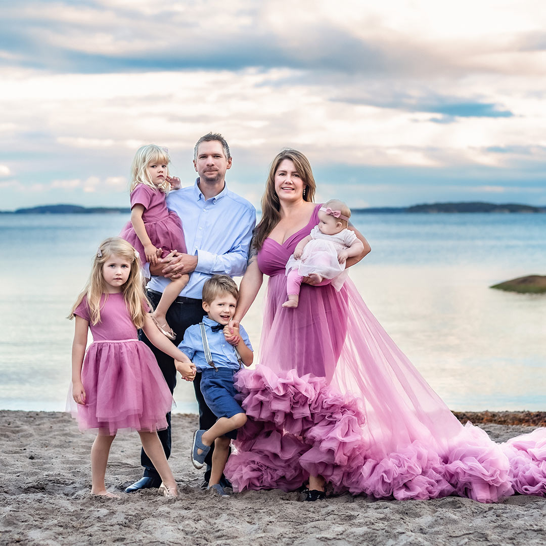 En finklädd familj i rosa toner står på en sandstrand. I bakgrunden syns havet och vacker himmel. Familjen tittar in i kameran.