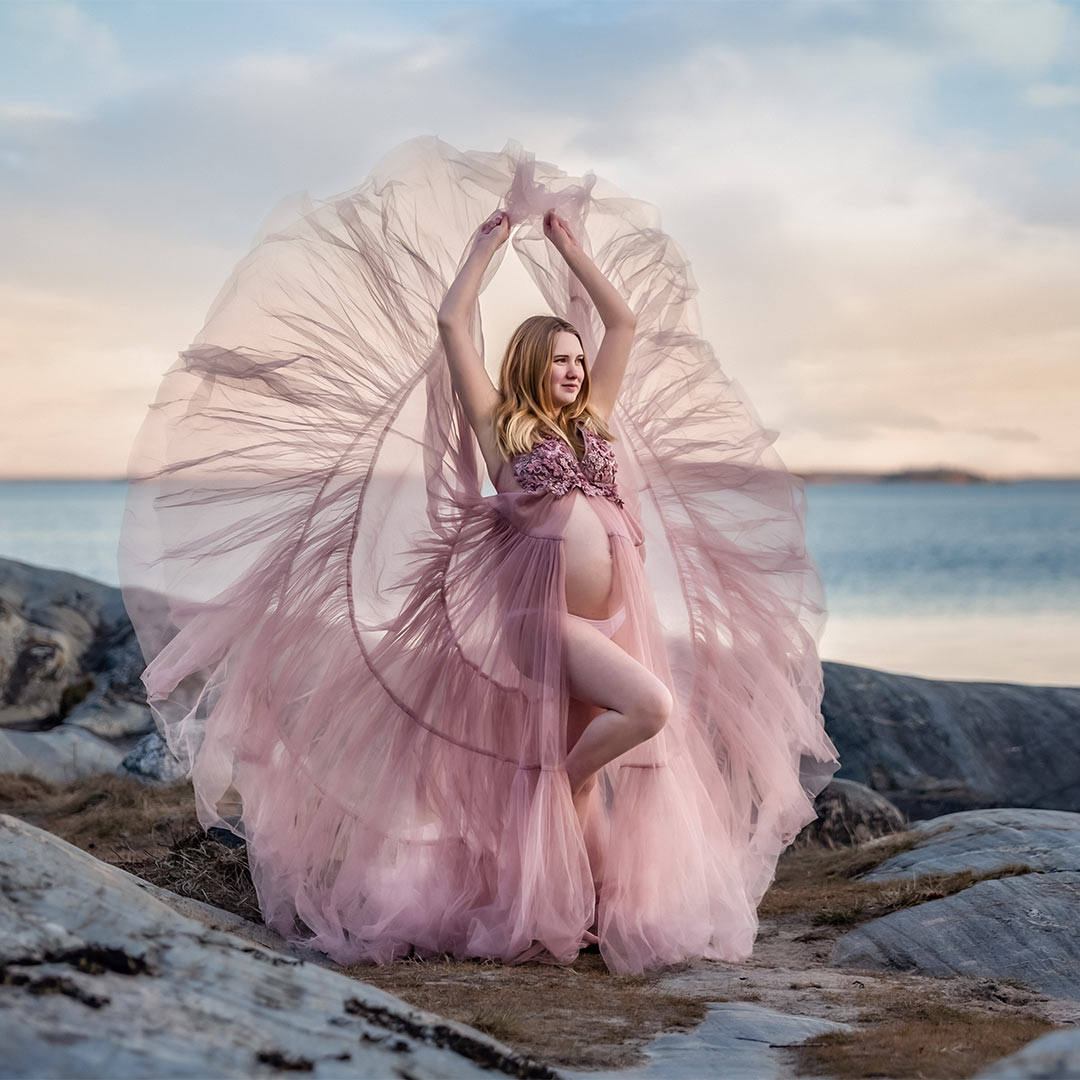 Gravidfotografering bland klipporna vid havet. Kvinnan har en rosa tyllklänning som flyger som vingar.