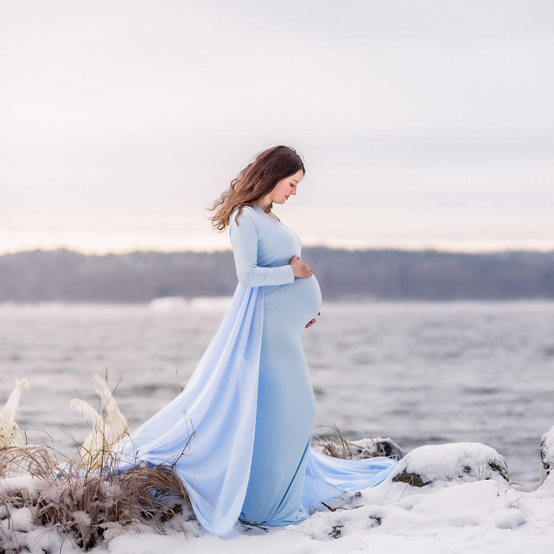 Gravidfotografering en riktigt kall dag när isen frusit gräset vid vattenkanten. Den gravida kvinnan har en blå klänning med släp och tittar ner på sin gravidmage.