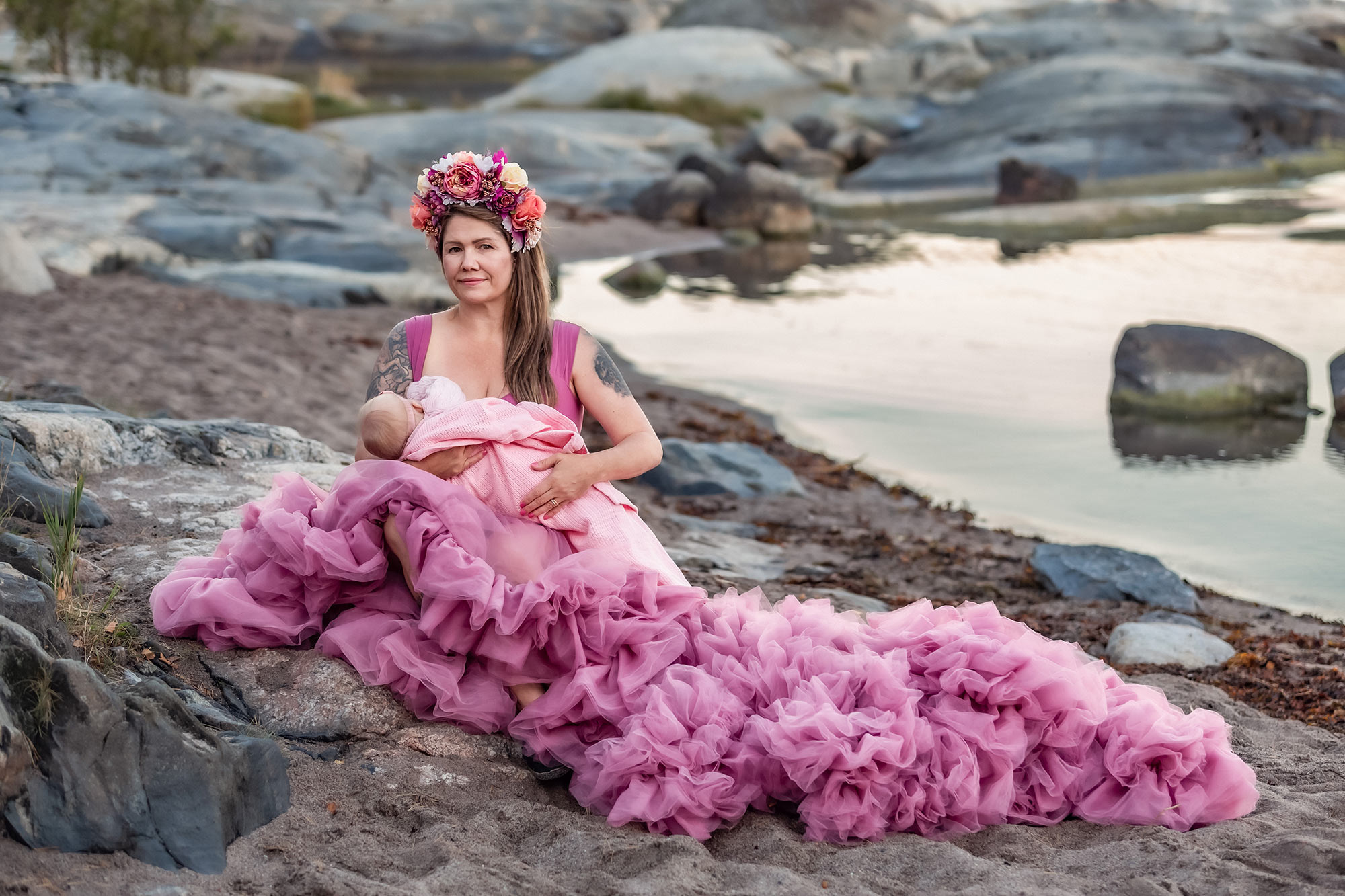 En mamma i rosa klänning sitter på stranden och ammar sitt lilla barn. I bakgrunden syns vatten och klippor.
