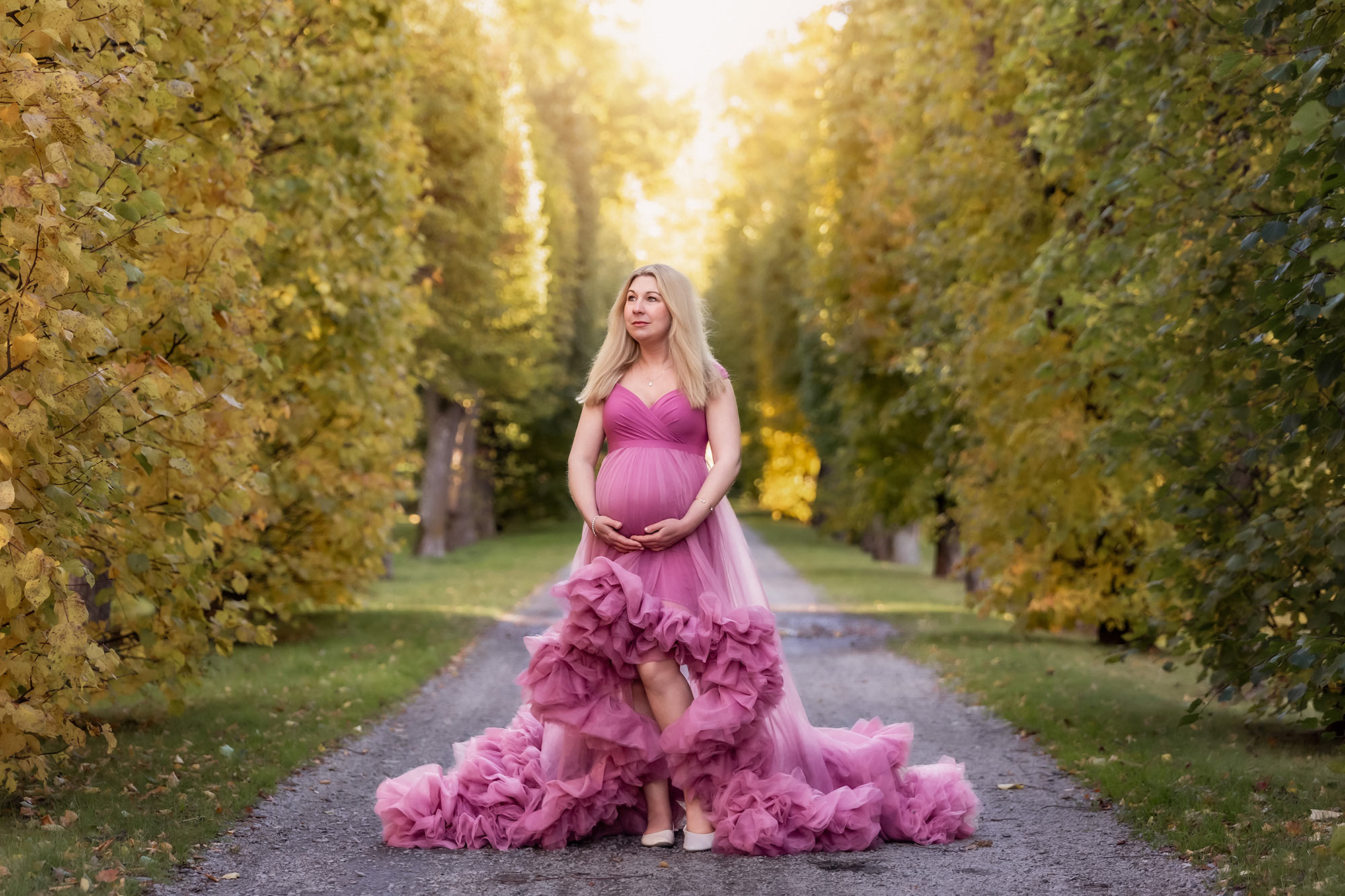 Gravidfotografering i en vacker höstfärgad allé. Den gravida kvinnan står på en grusgång och har på sig en starkrosa tyllklänning.