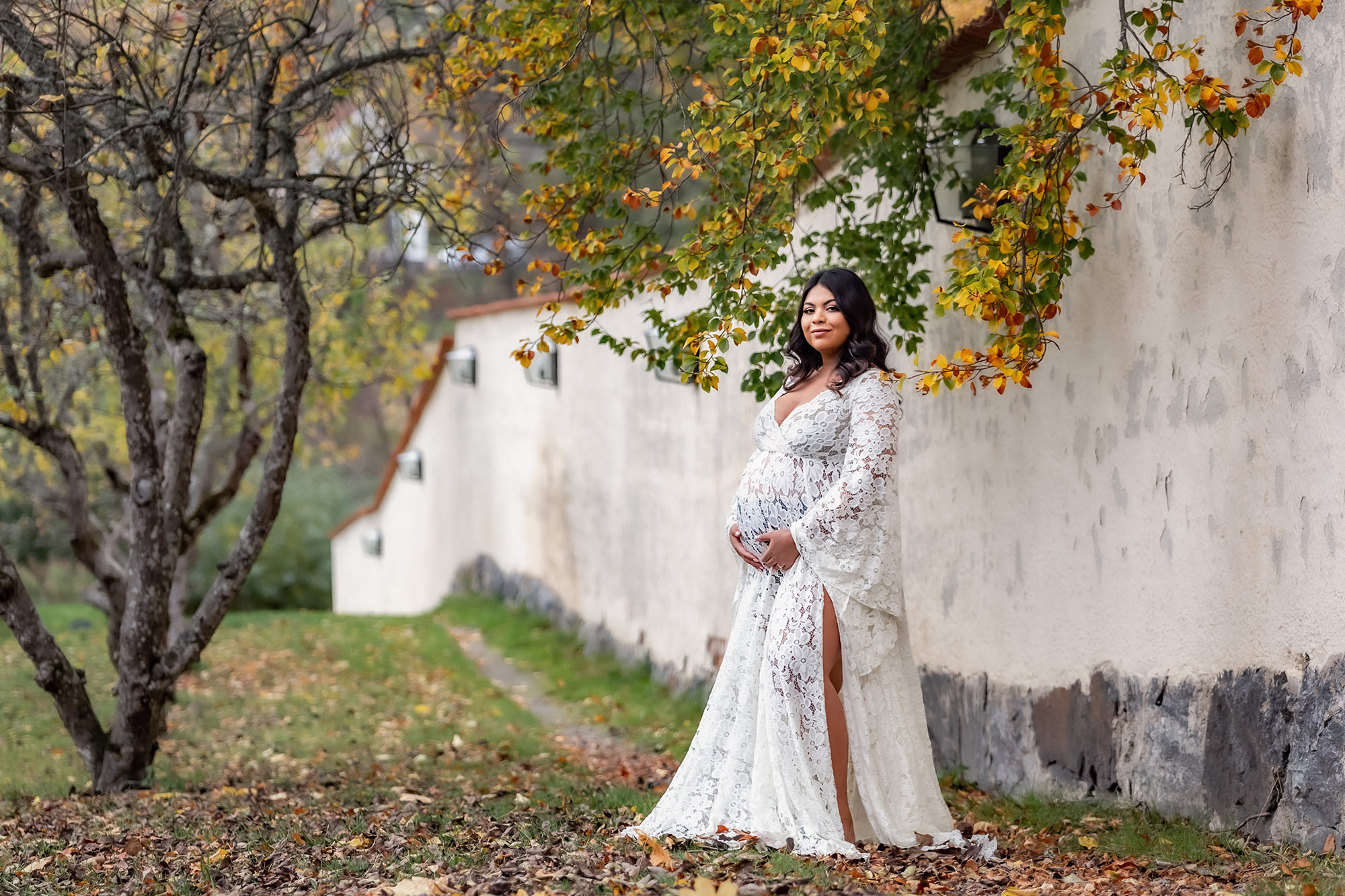 Gravidfotografering om hösten vid Tyresö slotts vackra vita mur. Den gravida kvinnan har en vit lång spetsklänning och runt kvinnan hänger grenarna ner med höstfärgade löv.