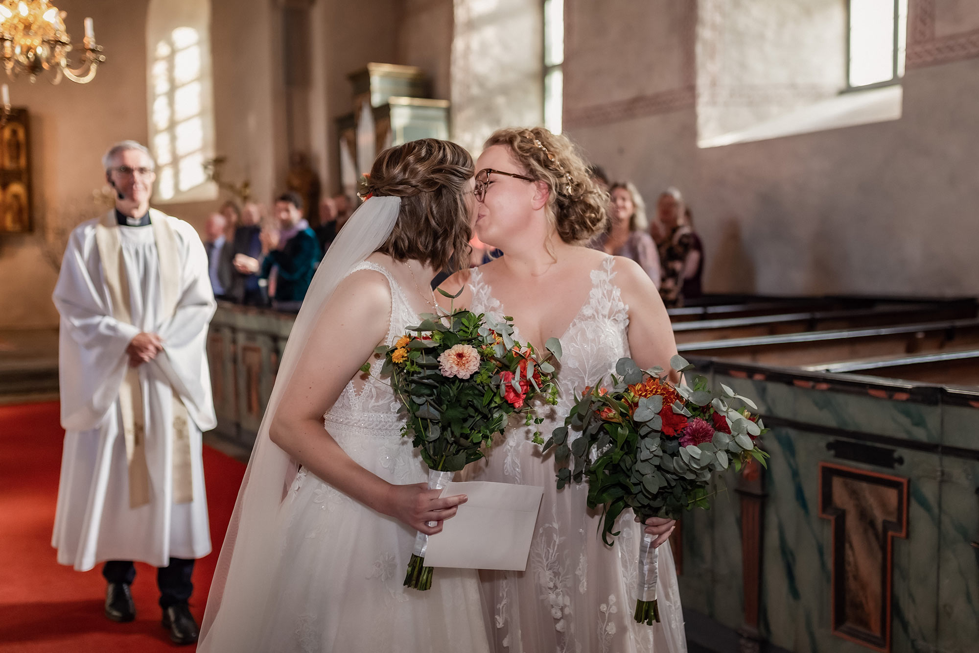 Ett nygift brudpar går i altargången och stannar till för en extra puss. I bakgrunden syns prästen och gästerna i Gamla Uppsala kyrka.
