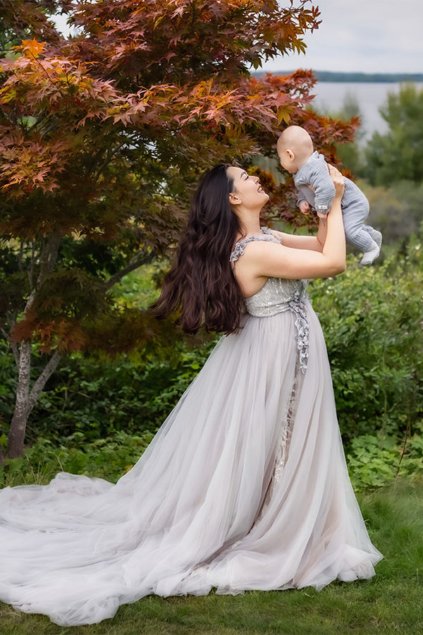 En vacker mamma i silverklänning står vid ett höstträd och håller upp sitt barn.