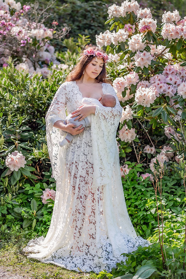 En mamma i vit spetsklänning ammar sitt barn stående bland blommande buskar.