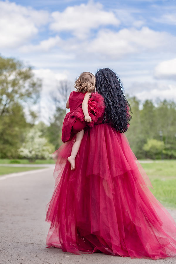 En mamma i röd klänning står på en stig en sommardag och håller i sin dotter som också har en röd klänning.