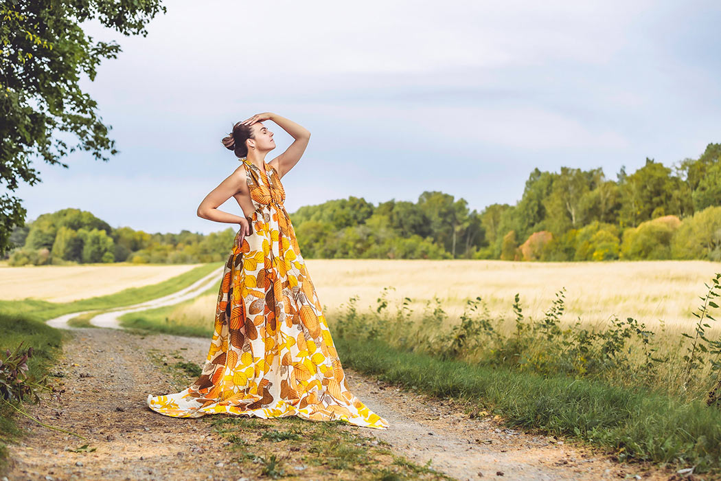 Kreativ fotografering i september i lantlig miljö. Kvinnan står på en grusväg bredvid åkrar och har på sig en klänning med stora höstlöv.