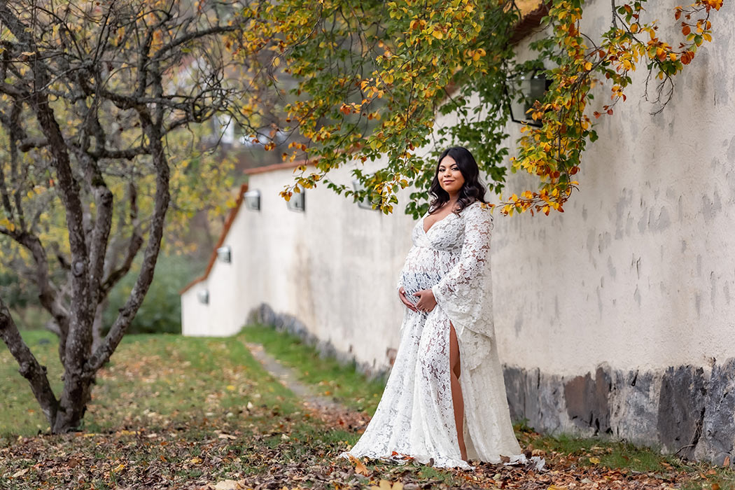 En gravid kvinna har på sig en vit spetsklänning och står framför en lång vit mur. Grenar hänger ner bakom kvinnan med höstlöv.