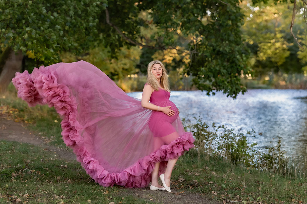 Gravidfotografering vid strandkanten i en slottsträdgård. Den gravida kvinnan har på sig en rosa tyllklänning med gigantiskt släp som flyger i vinden.