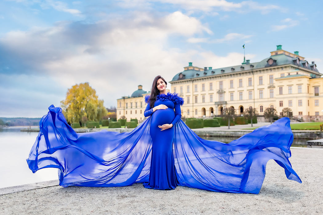 En gravid kvinna står framför Drottningholms slott i en blå klänning med ett enormt släp som flyger i vinden.