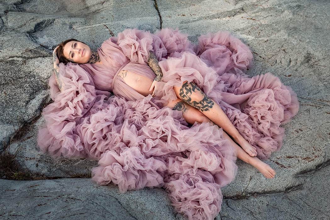 En vacker gravid kvinna ligger på en klippa. På sig har hon en rosa gravidklänning i tyll som är öppen fram.