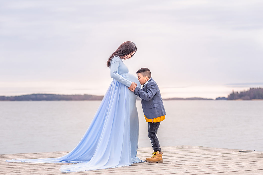 Gravidfotografering på en brygga vid havet. Kvinnan har en blå klänning och hennes son pussar hennes mage.