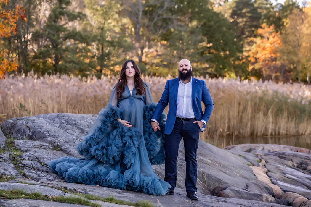 Ett par står hand i hand på en klippa vid vattnet. I bakgrunden syns vass och höstfärgade träd. Kvinnan är gravid och har en vacker blå gravidklänning.