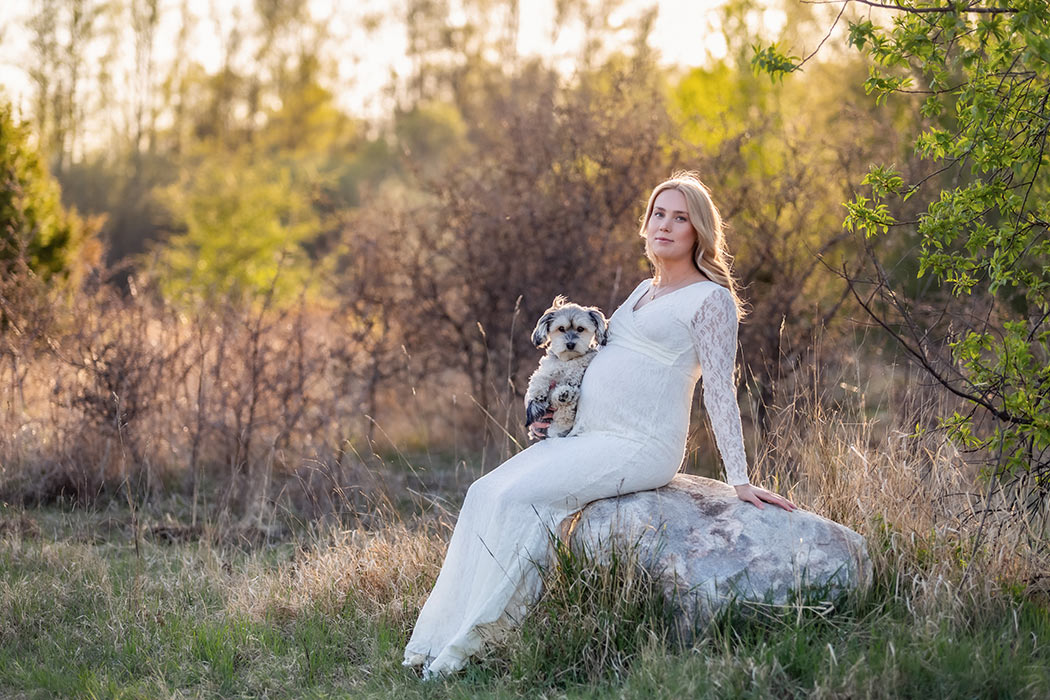 Gravid kvinna sitter på en stor sten bland äng och skog. I sin famn har hon sin lilla hund. Både hunden och kvinnan tittar in i gravidfotograf Linda Holmkratz kamera.