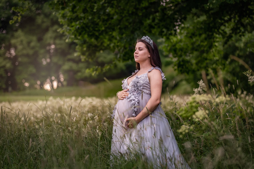 Gravidfotografering av en gravid kvinna som står i ett hav av gräs. I bakgrunden strålar solglimtar in och på sig har kvinnan en grå lyxig gravidklänning och en silverkrona som hon har lånat av gravidfotograf Linda Holmkratz för fotograferingen.
