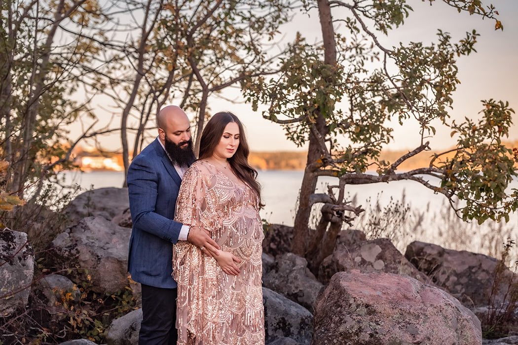 Ett par står nära varandra och håller kärleksfullt om kvinnans gravida mage. I bakgrunden syns stora stenbumlingar, träd och havet.