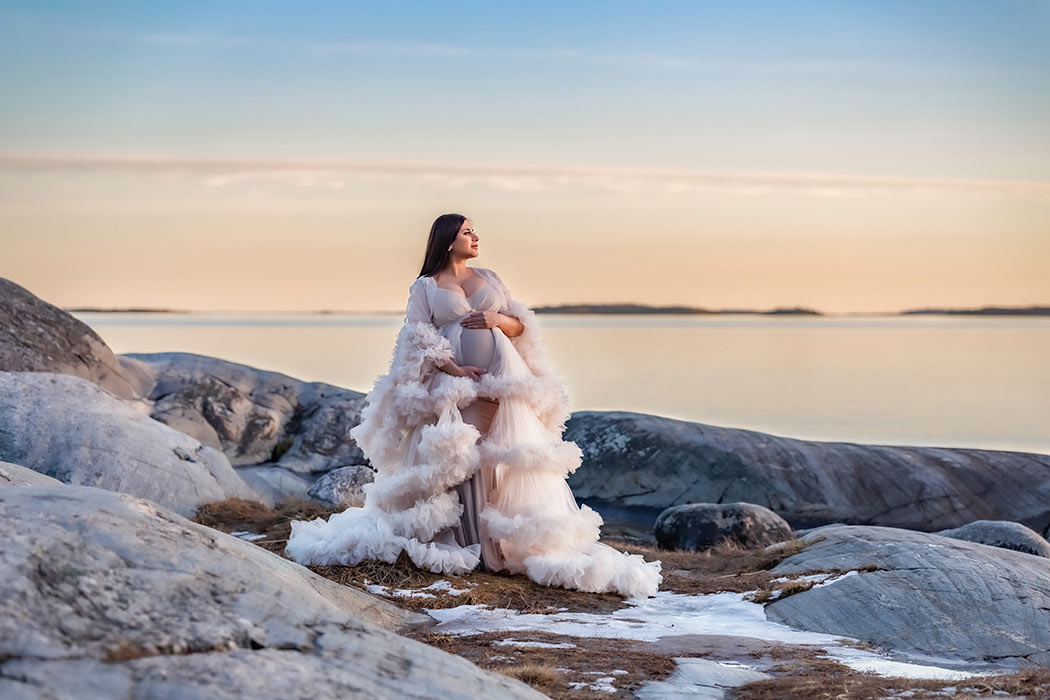 Gravidfotografering på klipporna vid havet i solnedgången. På marken ligger lite snö och is och kvinnan har en ljusbeige tyllklänning.