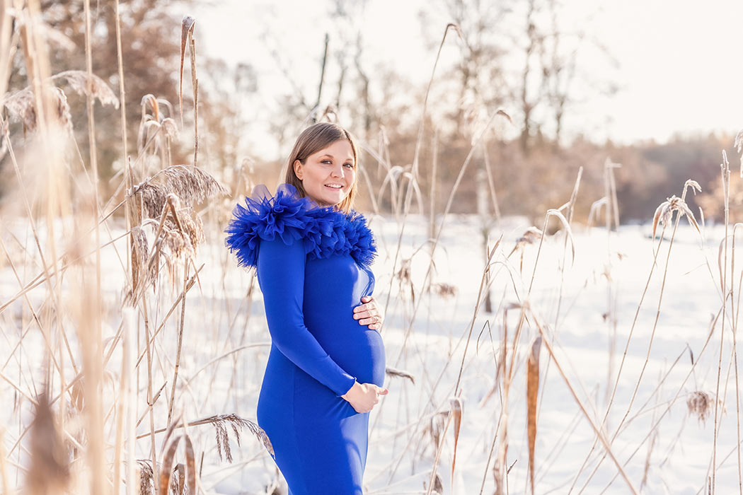 En gravid kvinna i en blå klänning står bland vassen en kall vinterdag i januari.