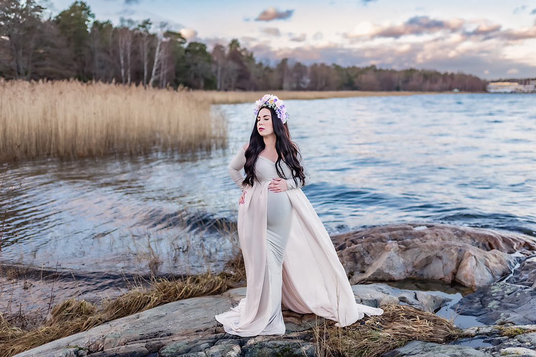 Gravidfotografering på klipporna vid vågande vatten. I bakgrunden syns vass och kala träd och den gravida kvinnan har en beige klänning med släp och en blomsterkrans.