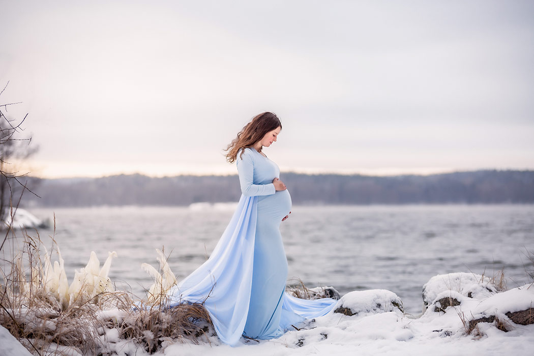 Gravidfotografering vid havet där vattenkantens strån har frusit till is. Snön ligger tjock på marken och i bakgrunden syns havet.