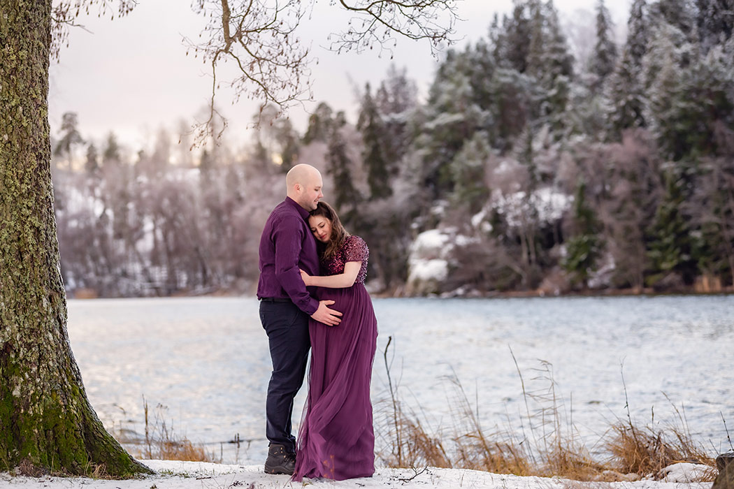 Parfotografering en kall vinterdag under ett stort träd. Paret är klädda i lila och kvinnan är gravid. På marken ligger snö och i bakgrunden syns ett porlande vatten och snötäckta granar.