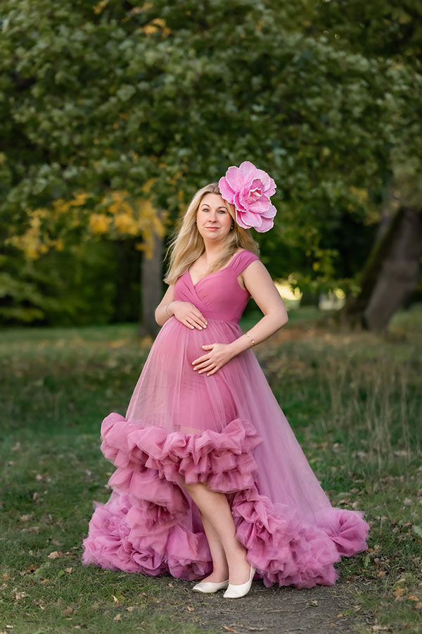 Gravidfotografering bland gräs och stora träd. Den gravida kvinnan har på sig en starkt rosa tyllklänning och på huvudet har hon en stor rosa blomma.