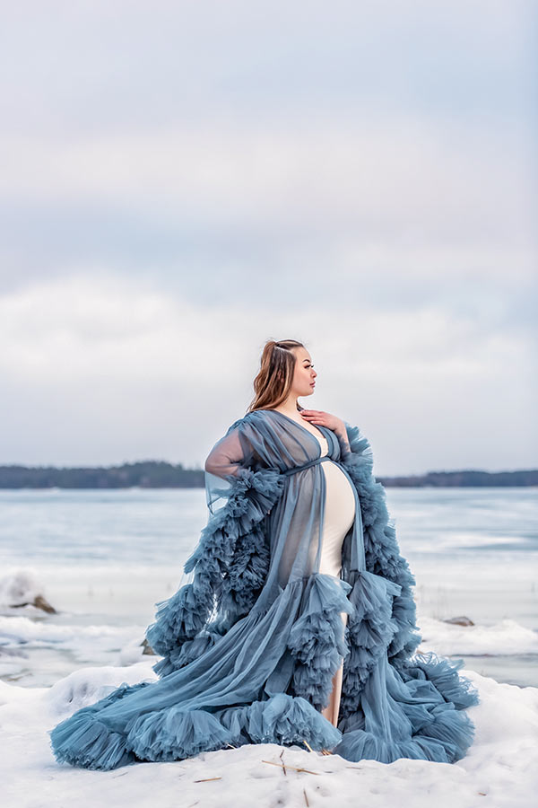 Gravidfotografering på vintern vid ett istäckt hav. Den gravida kvinnan har på sig en blå tyllklänning.