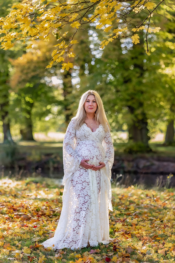 Gravidfotografering om hösten. Den gravida kvinnan har på sig en vit spetsklänning och står under en gren med gula lönnlöv. På marken ligger fullt med höstlöv och i bakgrunden syns träd och en liten å.