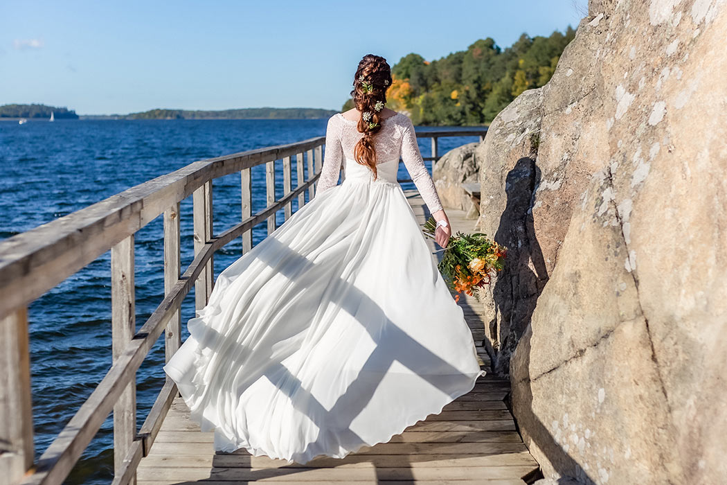 En brud klädd i bröllopsklänning och bärandes en brudbukett går på en bryggpromenad längs Mälaren i Hässelby vid Riddersvik. Brudklänningen fladdrar i vinden och i bakgrunden syns vatten med segelbåt och höstiga träd.