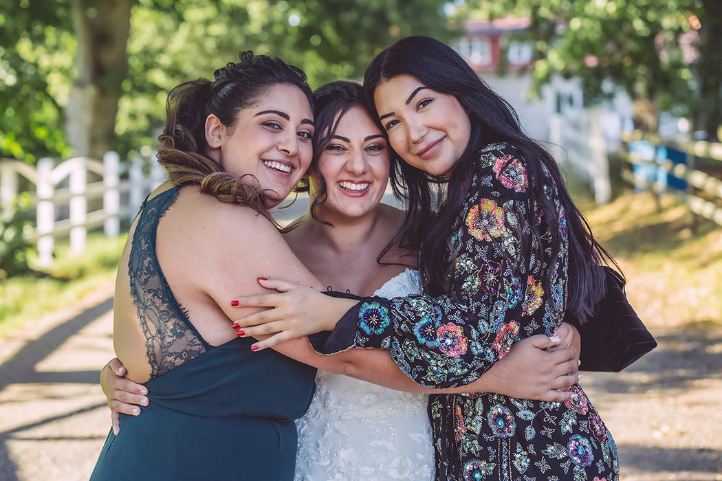 En nygift brud och hennes två vänner står på en grusgång vid Ekeby loge och tittar in i bröllopsfotografens kamera och skrattar.