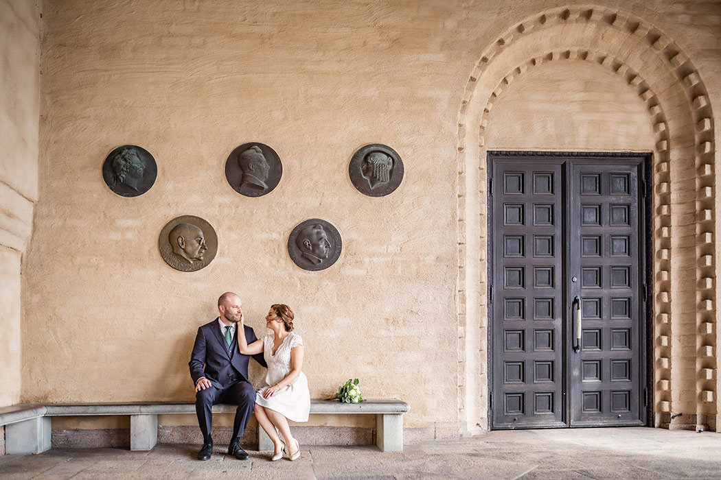 Brudpar sitter på en stenbänk vid en stor pampig dörr vid ingången till Stockholms stadshus. Bruden klappar brudgummen på kinden och bredvid paret ligger brudbuketten.
