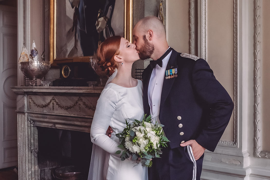 Ett nygift par pussas vid spiselkransen i ett pampigt rum. Kvinnan har en vit klänning och mannen har militär klädsel.