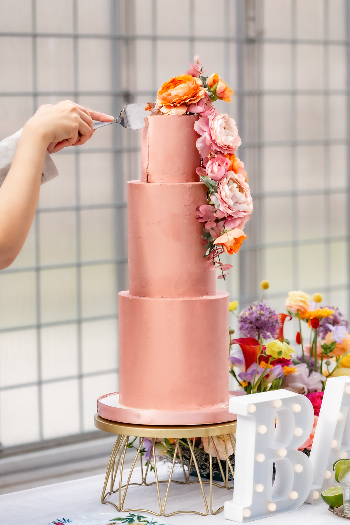 Två händer håller i en tårtspade som skär en bit av en hög, rosa bröllopstårta. Tårtan är dekorerad med levande blommor.