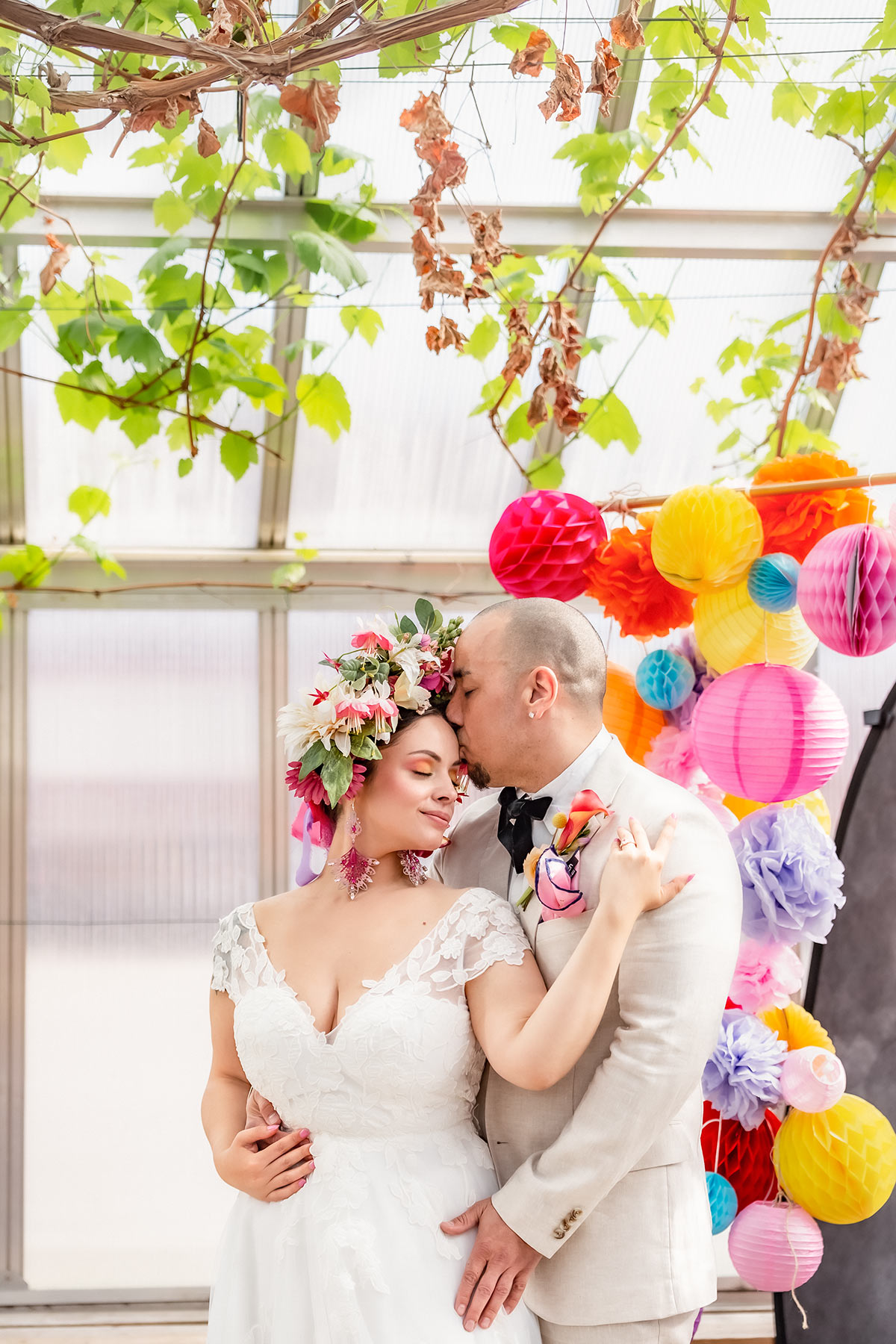 Ett brudpar håller om varandra framför färgglada dekorationen. De står i ett växthus och i taket hänger vinrankor. Brudgummen pussar brudens panna.